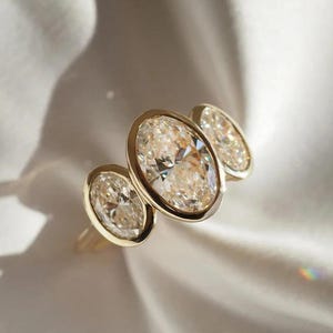 May include: A gold ring featuring three oval-cut diamonds. The diamonds are set in a bezel setting, and the ring is displayed on a white, silky fabric. The diamonds have a warm, champagne color and sparkle in the light.
