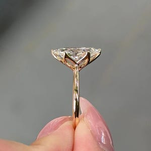 May include: A gold ring featuring a large, clear, pear-shaped gemstone. The gemstone is held in place by delicate prongs, and the band is a thin, polished gold. The ring is held up against a soft gray background.