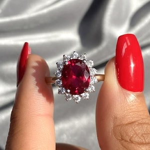 May include: A gold ring featuring a large, oval-cut, deep red gemstone surrounded by a halo of smaller, clear gemstones. The ring is held up by a hand with red nail polish against a silver background.