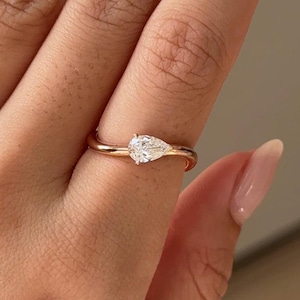 May include: A gold ring with a pear-shaped diamond. The ring is set on a slender band. The ring is shown on a hand with manicured nails.