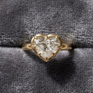 May include: A gold ring with a large, heart-shaped diamond. The diamond is clear and faceted, set in a gold bezel. The ring is on a gray, textured surface.