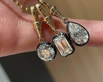 May include: Three gold pendant necklaces with different diamond shapes: a round, an emerald cut, and a pear shape. The diamonds are set in black bezels and hang from gold chains. The pendants are displayed against a neutral background.