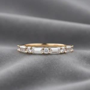 Può includere: Un anello a fascia in oro con diamanti taglio baguette e rotondi alternati. L'anello presenta un motivo continuo di pietre preziose rettangolari e circolari, creando un design scintillante ed elegante. L'anello è posto su uno sfondo grigio.