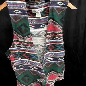 May include: A sleeveless vest featuring a multi-colored woven pattern in shades of green, red, blue, brown, and white. The garment has a single toggle closure at the bottom front and a label at the neckline that reads STAMPEDE MADE IN U.S.A.