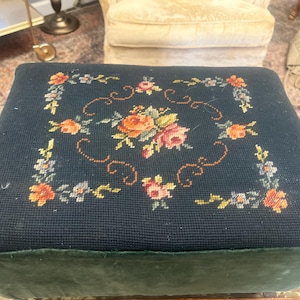 May include: A dark blue needlepoint footstool with a floral design. The top features a central arrangement of roses in shades of orange, pink, and yellow, surrounded by smaller flowers and green leaves. The sides are a dark green velvet.