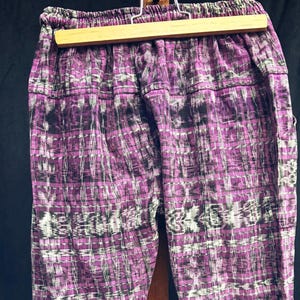 May include: A pair of purple and black trousers featuring an intricate ikat-style woven pattern with white accents. The pants have an elasticized waistband and are displayed hanging from a wooden hanger against a dark background.