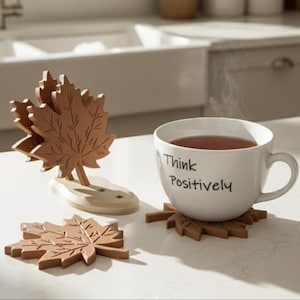 Könnte beinhalten: Eine weiße Keramiktasse mit dem in Schwarz aufgedruckten Text Think Positively, die ein dampfendes Getränk enthält. Die Tasse steht auf einem braunen Untersetzer in Ahornblattform. Daneben befindet sich ein passender Halter mit weiteren Untersetzern.