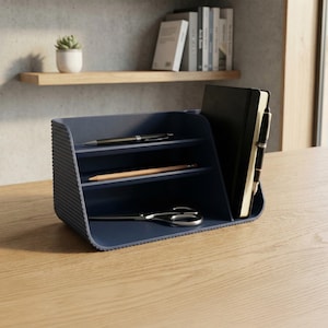 May include: A dark blue desk organizer with a ribbed exterior featuring three horizontal shelves and a vertical compartment. The organizer holds a black notebook, a black pen, a wooden pencil, a pair of metal scissors, and a fountain pen.