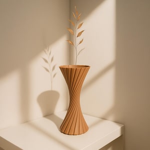 May include: A terracotta-colored vase with a twisted, ribbed design. The vase is holding a dried plant with small, brown leaves. The vase is on a white surface, with a shadow of the plant on the wall.