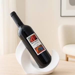 May include: A dark glass wine bottle with a black cap and a rectangular label featuring abstract red, orange, and blue shapes, resting at an angle in a white, curved, wave-shaped holder with a textured matte finish.