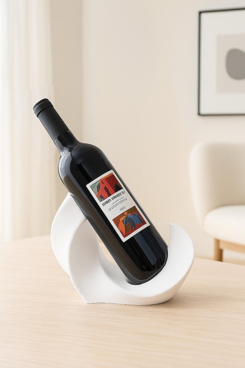 May include: A dark glass wine bottle with a black cap and a rectangular label featuring abstract red, orange, and blue shapes, resting at an angle in a white, curved, wave-shaped holder with a textured matte finish.