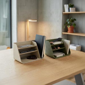 May include: Two desk organizers with a ribbed exterior texture, one in beige and one in olive green. Each organizer features three tiered shelves and a vertical slot for notebooks, holding pens, scissors, and small journals on a wooden surface.