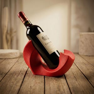 May include: A red, crescent-shaped wine bottle holder with a textured surface, cradling a dark glass wine bottle with a cream-colored label that reads CANWE-RO. The bottle is positioned at an angle, resting securely within the curved structure.