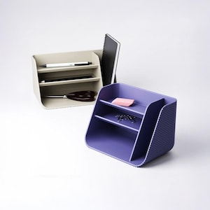 May include: Two desk organizers with a ribbed exterior texture. One organizer is beige and holds a white pen, a black pen, a black notebook, and black scissors. The other organizer is purple and holds a pink eraser and silver paper clips.