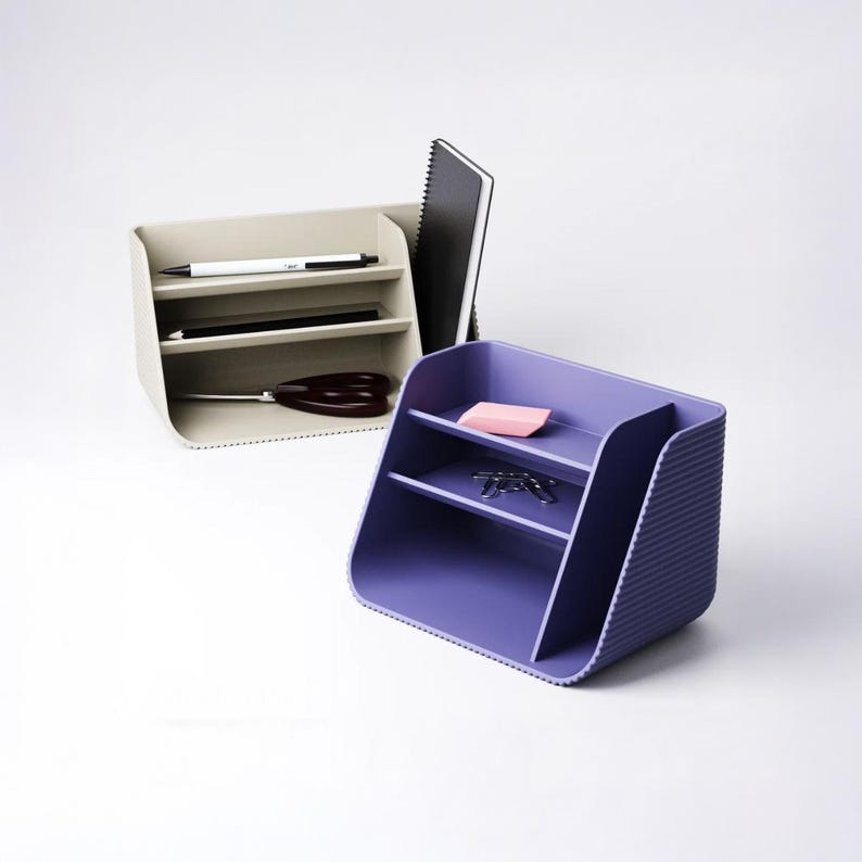 May include: Two desk organizers with a ribbed exterior texture. One organizer is beige and holds a white pen, a black pen, a black notebook, and black scissors. The other organizer is purple and holds a pink eraser and silver paper clips.