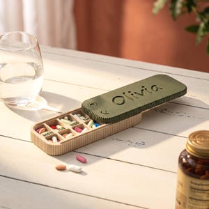 Puede incluir: Un pastillero rectangular con tapa deslizante de color verde oliva que tiene el nombre Olivia grabado en relieve. La base es de color beige claro con lados acanalados y múltiples compartimentos etiquetados con los días de la semana, con varias pastillas de colores.