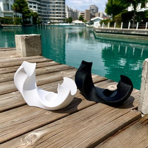 May include: Two abstract sculptural objects, one in matte white and one in matte black, featuring curved, organic forms with a textured surface. Each piece has a central circular indentation and sweeping, wave-like edges that rise to points.