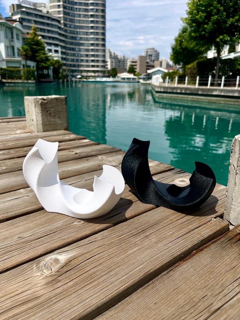May include: Two abstract sculptural objects, one in matte white and one in matte black, featuring curved, organic forms with a textured surface. Each piece has a central circular indentation and sweeping, wave-like edges that rise to points.