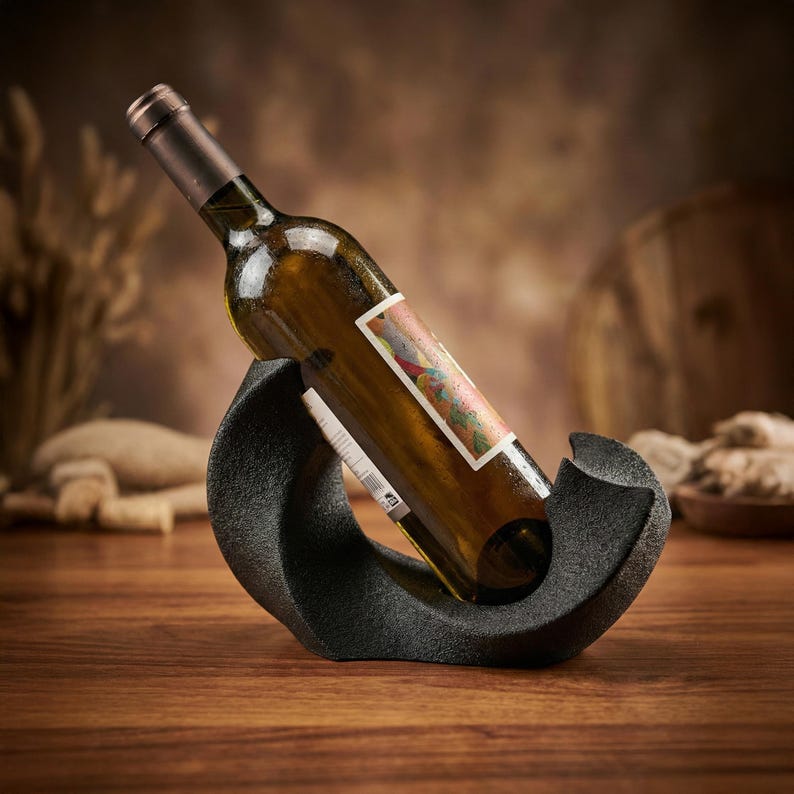 May include: A dark gray, textured, curved wine bottle holder cradling a glass bottle of white wine. The bottle is positioned at an angle within the holder, featuring a rectangular label with colorful abstract patterns and visible text.