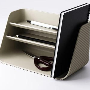 May include: A beige desk organizer with a ribbed exterior, featuring two horizontal shelves and a vertical slot. The organizer holds a white pen, a black pencil, a pair of dark brown scissors, and a black spiral-bound notebook standing upright.