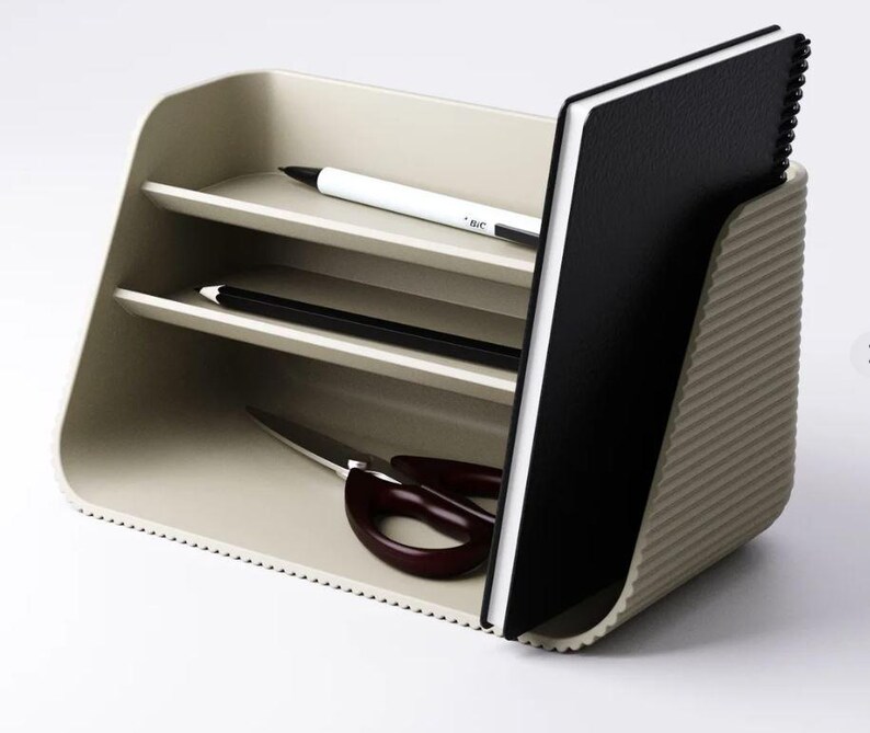 May include: A beige desk organizer with a ribbed exterior, featuring two horizontal shelves and a vertical slot. The organizer holds a white pen, a black pencil, a pair of dark brown scissors, and a black spiral-bound notebook standing upright.
