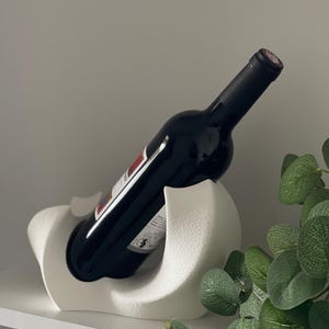 May include: A white wine bottle holder with a textured, matte finish and a curved, sculptural design. The holder supports a dark glass wine bottle at an angle, cradling the base and body within its smooth, contoured, and rounded white structure.