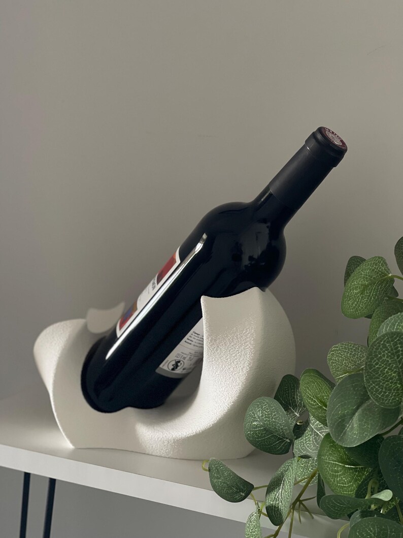 May include: A white wine bottle holder with a textured, matte finish and a curved, sculptural design. The holder supports a dark glass wine bottle at an angle, cradling the base and body within its smooth, contoured, and rounded white structure.