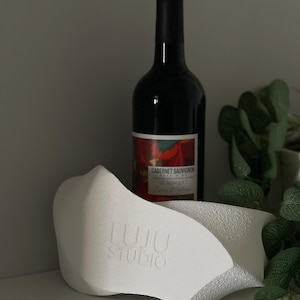 May include: A white, textured, sculptural wine bottle holder with an irregular, organic shape and the embossed text LUJU STUDIO on the front, positioned in front of a dark glass bottle of Cabernet Sauvignon wine with a red and white label.