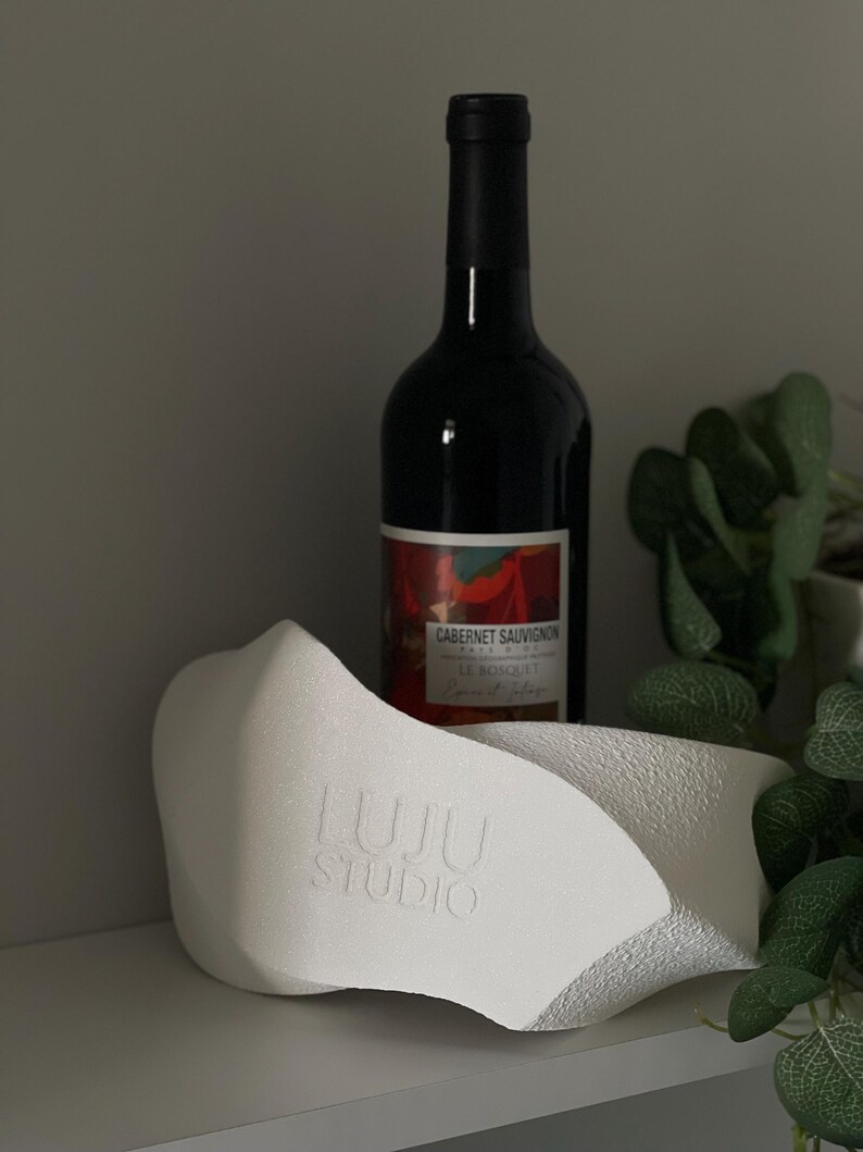 May include: A white, textured, sculptural wine bottle holder with an irregular, organic shape and the embossed text LUJU STUDIO on the front, positioned in front of a dark glass bottle of Cabernet Sauvignon wine with a red and white label.