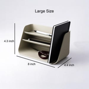 May include: A beige desk organizer with a ribbed side panel, featuring two horizontal shelves and a vertical slot. It holds a black notebook, two pens, and scissors. Text indicates Large Size, 4.3 inches high, 8 inches wide, and 4.4 inches deep.