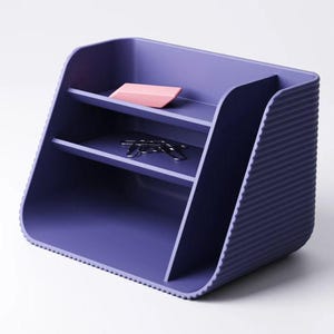 May include: A purple desk organizer with a ribbed exterior texture. The unit features two horizontal shelves and a tall vertical compartment. A pink rectangular eraser sits on the top shelf, and several silver metal paper clips rest on the middle shelf.