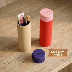 Op de afbeelding: Drie cilindrische reiscontainers in beige, rood en paars. De beige container bevat een tandenborstel en tandpasta. De rode container heeft een roze deksel met de naam "Emma". De paarse deksel is gegraveerd met de naam "James".