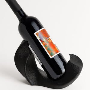 May include: A black wine bottle with a colorful abstract label featuring orange, red, and purple tones rests at an angle within a curved, textured black wine rack. The rack has a matte finish and a smooth, sweeping shape that supports the bottle.