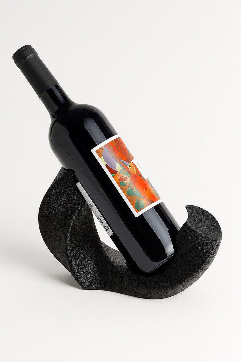 May include: A black wine bottle with a colorful abstract label featuring orange, red, and purple tones rests at an angle within a curved, textured black wine rack. The rack has a matte finish and a smooth, sweeping shape that supports the bottle.