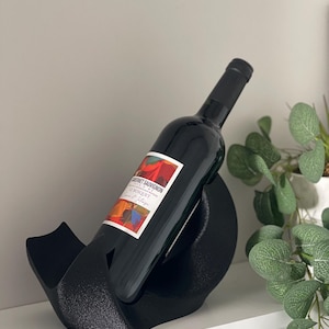 May include: A black, textured, curved wine bottle holder cradling a dark glass bottle of Cabernet Sauvignon. The bottle features a white label with red, orange, and blue abstract graphic patterns and the text Le Bosquet printed in black.
