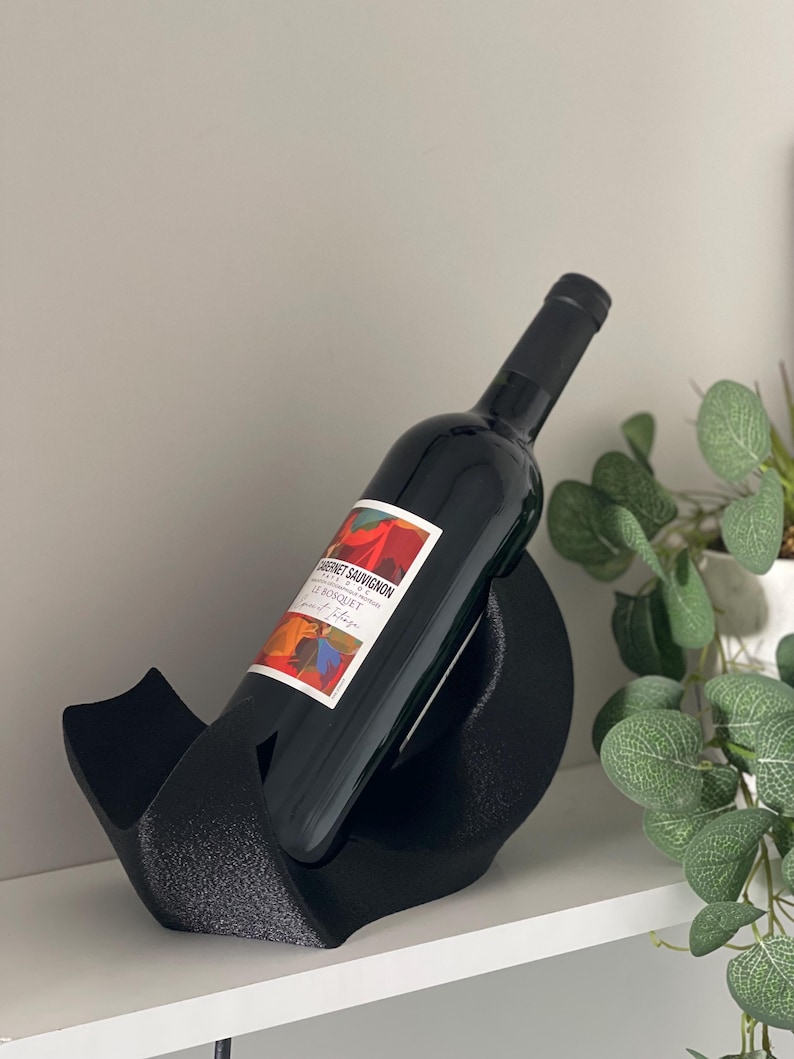 May include: A black, textured, curved wine bottle holder cradling a dark glass bottle of Cabernet Sauvignon. The bottle features a white label with red, orange, and blue abstract graphic patterns and the text Le Bosquet printed in black.