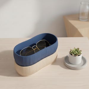 May include: A blue and cream oval storage container with a ribbed design, containing gold-rimmed sunglasses. A small succulent in a white pot sits on a grey coaster. The items are on a light wood surface.