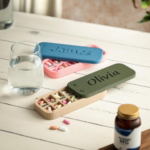 Könnte beinhalten: Zwei rechteckige Pillendosen mit Schiebedeckeln, eine rosa mit blauem Deckel beschriftet mit James und eine beige mit olivgrünem Deckel beschriftet mit Olivia. Jeder Behälter hat sieben Fächer mit Wochentagen und ist mit verschiedenen Pillen gefüllt.