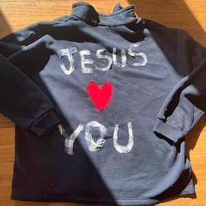 Puede incluir: Sudadera azul marino con las palabras "JESÚS TE AMA" aplicadas en tela blanca. Un corazón rojo está centrado entre las palabras. La sudadera tiene cuello alto y mangas largas.