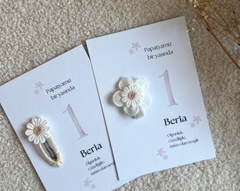 Può includere: Due fermagli per capelli bianchi con motivi a margherita ricamati e centri rosa chiaro, fissati su cartoncini bianchi con il testo Papatyamız bir yaşında, il numero 1, il nome Beria e altro testo in turco.