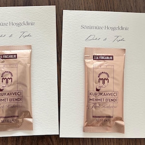 Peut inclure: Deux cartes rectangulaires blanc cassé avec le texte "Sözümüze Hoşgeldiniz" et des noms manuscrits. Chaque carte porte un sachet individuel en cuivre métallique de Kurukahveci Mehmet Efendi Türk Kahvesi, marqué "TEK FİNCANLIK" et "NET 6 g".
