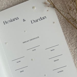 Może przedstawiać: Biały certyfikat Nikkah z imionami Besiana & Dardan w czarnym tekście. Dokument zawiera linie na datę, imama, mahr, pana młodego, pannę młodą i świadk&oacute;w, wraz z cytatem And We created you in pairs. An-Naba 78:8.