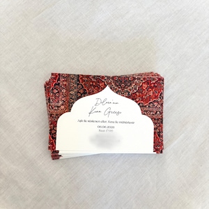 May include: A stack of invitation cards with a red and black patterned design, resembling a rug. Each card has a white cutout with text in a script font. The cards are stacked on a white linen surface.