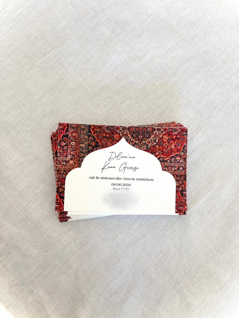 May include: A stack of invitation cards with a red and black patterned design, resembling a rug. Each card has a white cutout with text in a script font. The cards are stacked on a white linen surface.
