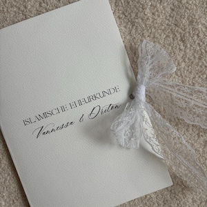 Pode incluir: Um cart&atilde;o branco com o texto "ISLAMISCHE EHRURKUNDE" e os nomes "Vanessa & Driton" em escrita elegante. Uma delicada fita de renda branca est&aacute; amarrada ao redor do cart&atilde;o.