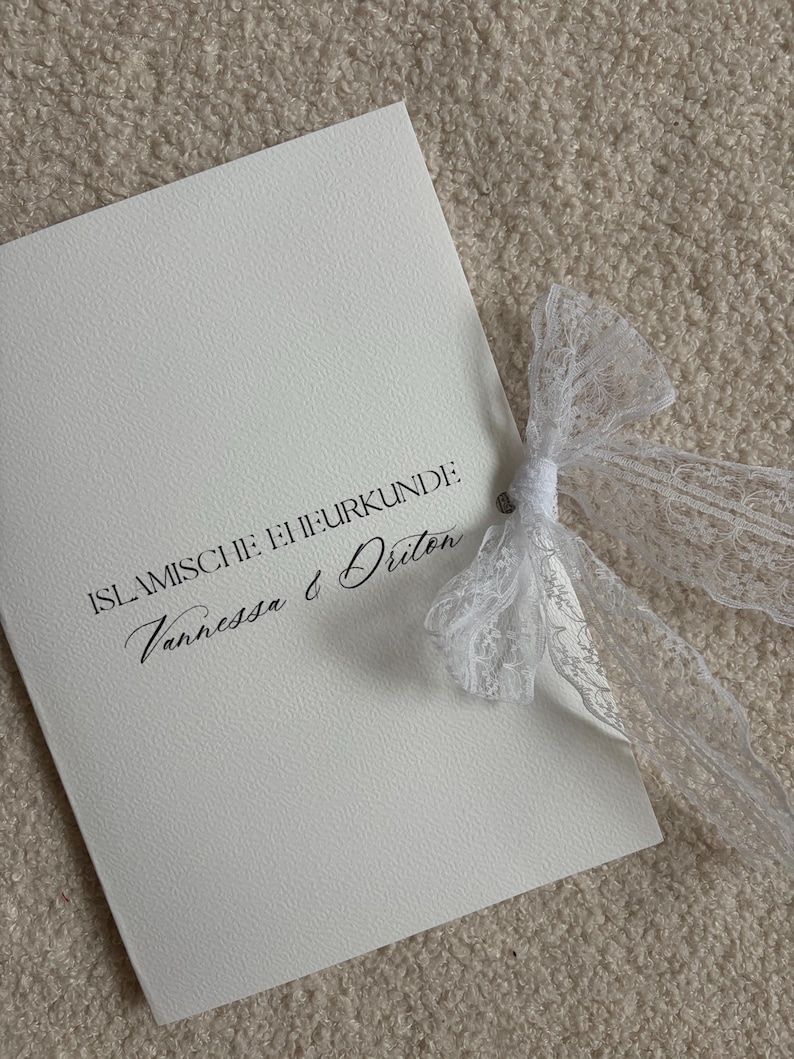 Pode incluir: Um cart&atilde;o branco com o texto "ISLAMISCHE EHRURKUNDE" e os nomes "Vanessa & Driton" em escrita elegante. Uma delicada fita de renda branca est&aacute; amarrada ao redor do cart&atilde;o.