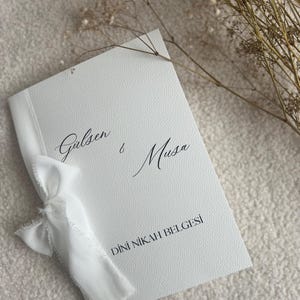 Op de afbeelding: Een wit gestructureerd boekje met een gerafeld wit lint aan de linkerkant. De omslag bevat de in zwart gedrukte namen Galsen & Musa en de tekst DİNİ NİKAH BELGESİ in een schreeflettertype, op een zachte, lichtgekleurde ondergrond.