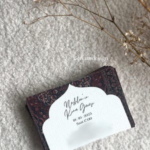 May include: A stack of invitation cards with a patterned burgundy and black design. The cards have a white cutout with calligraphy text and the date October 18, 2025, and time 5:00 PM. Dried flowers are in the background.