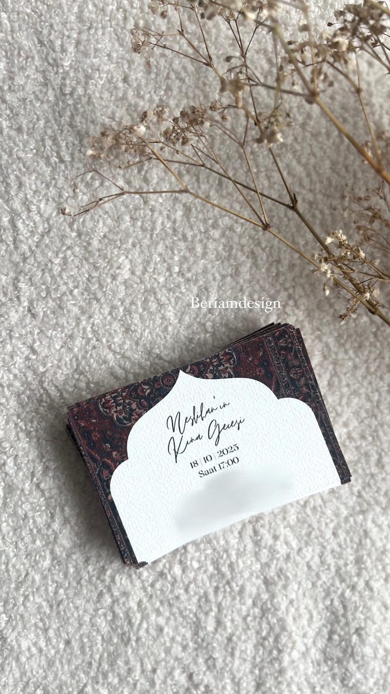 May include: A stack of invitation cards with a patterned burgundy and black design. The cards have a white cutout with calligraphy text and the date October 18, 2025, and time 5:00 PM. Dried flowers are in the background.