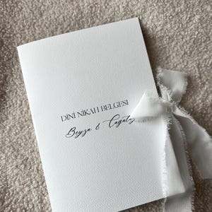 Pode incluir: Um convite branco com o texto "DINI NIKAH BELGESI" e os nomes "Beyza & Cagatay" em escrita cursiva. Uma fita branca est&aacute; amarrada ao redor do convite.